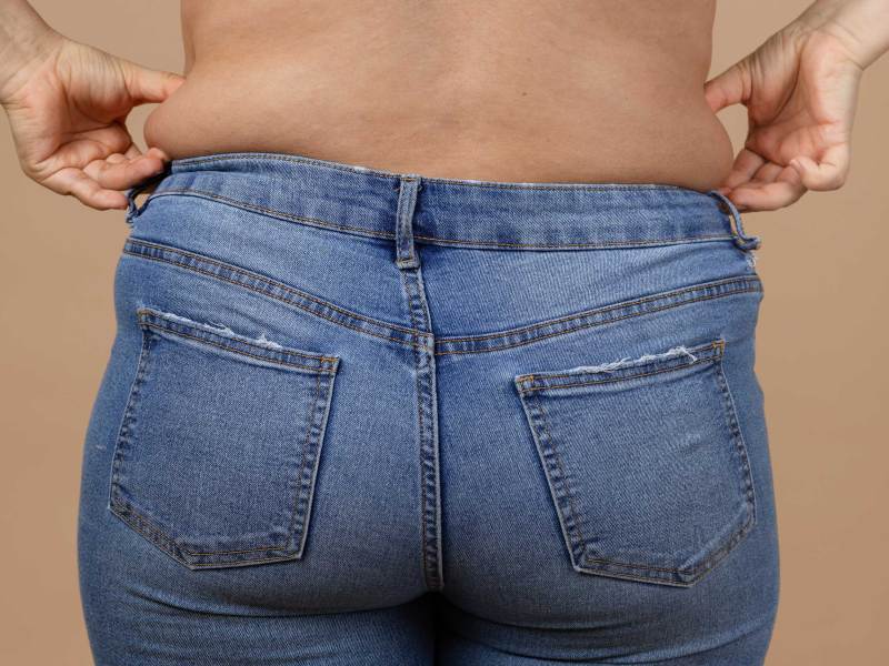 Nahaufnahme einer jungen Frau mit &uuml;bergewichtiger Taille, die eine blaue Jeans tr&auml;gt und mit den H&auml;nden H&uuml;ftspeck h&auml;lt.