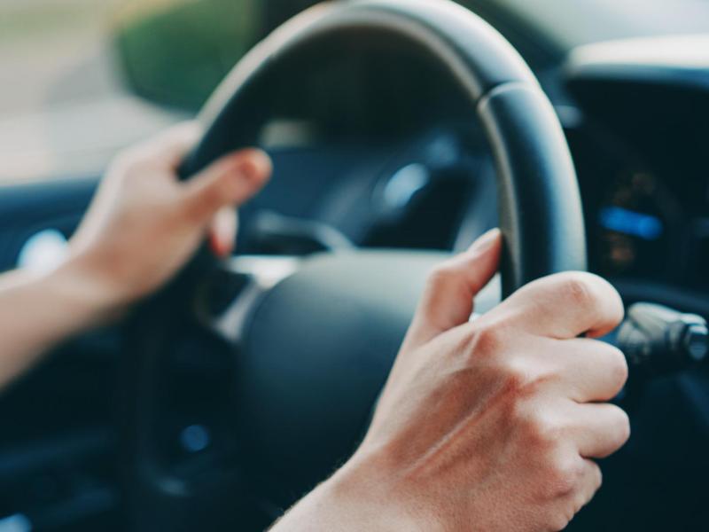 Nahaufnahme zweier Hände am Lenkrad eines Autos, mit unscharfem Armaturenbrett und Straßenhintergrund im Abendlicht.