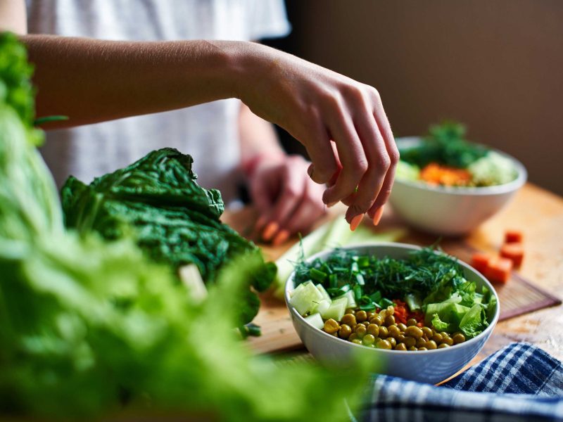 Weibliche Hand gibt grüne Zwiebeln in eine Schüssel mit grünen Erbsen, Gurken, Karotten, Salat und Dill, die auf einem Tisch stehen
