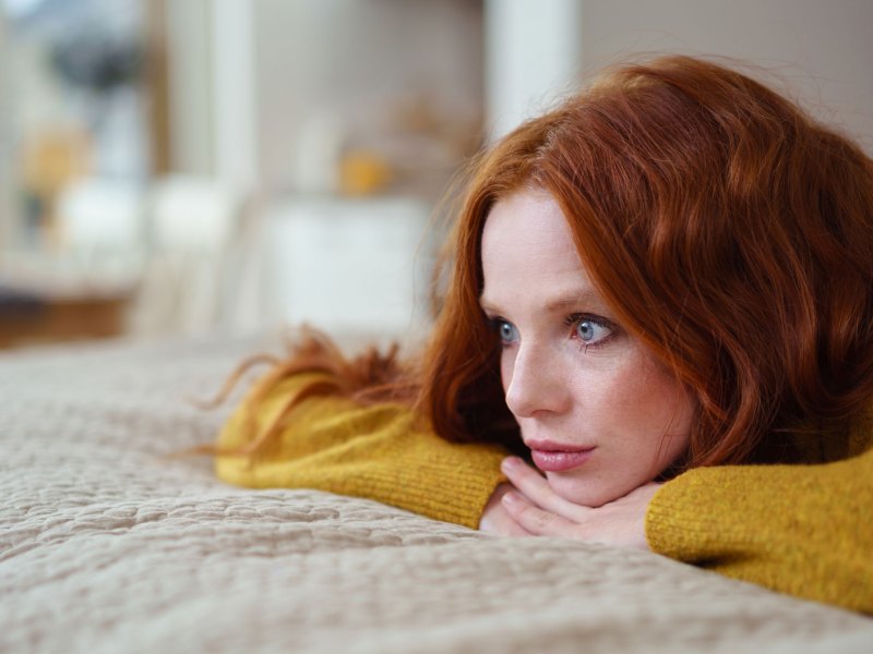 Frau mit roten Haaren und gelbem Pullover liegt auf dem Bett und blickt ernst.