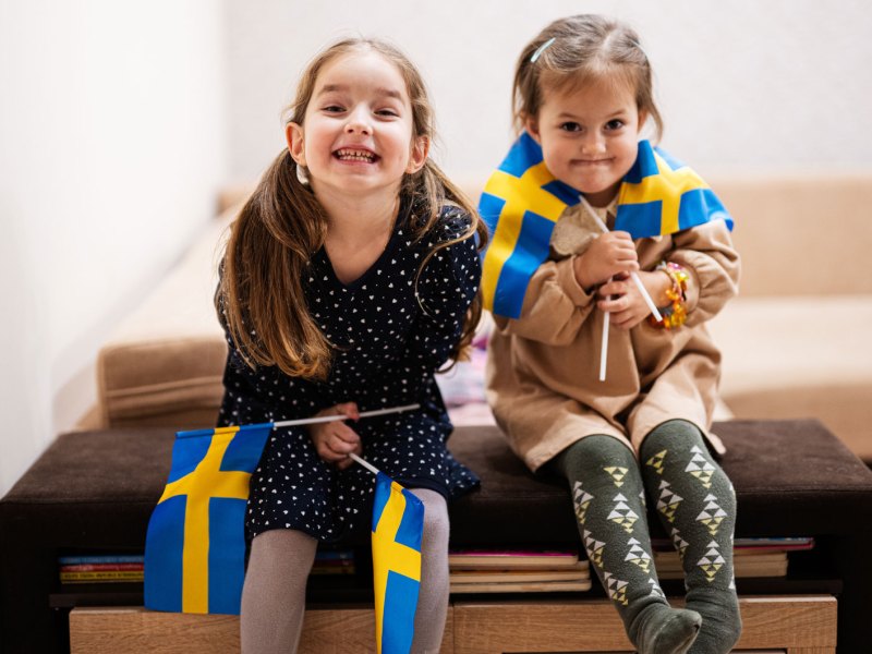 Zwei kleine M&auml;dchen sitzen auf einer gro&szlig;en Kiste und halten je zwei schwedische Flaggen in ihren H&auml;nden.
