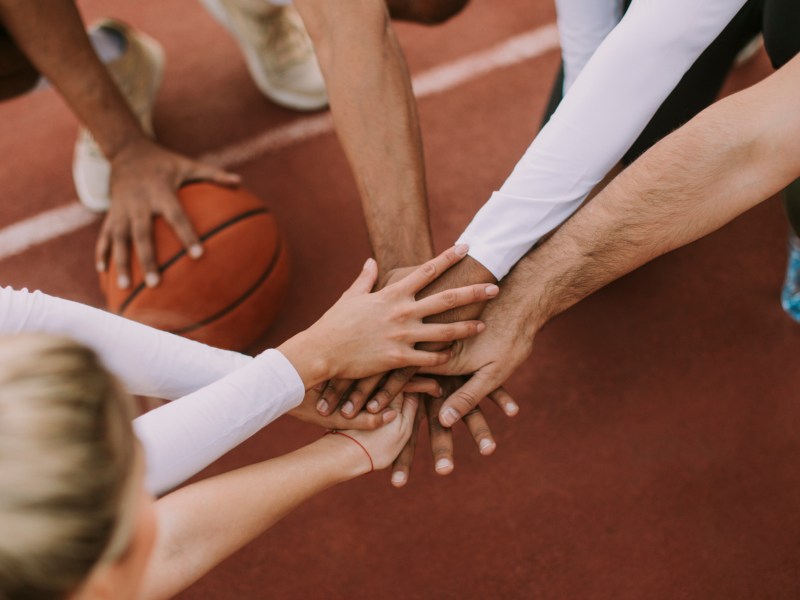 Menschen hocken auf einem Basketballplatz im Kreis und legen in der Mitte die H&auml;nde zusammen.