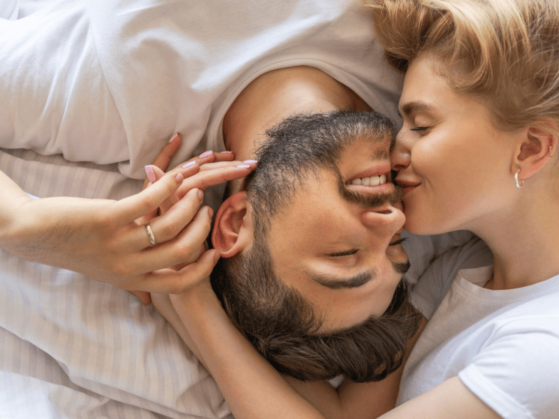 Frau neben Mann im Bett, die ihn auf die Wange k&uuml;sst.