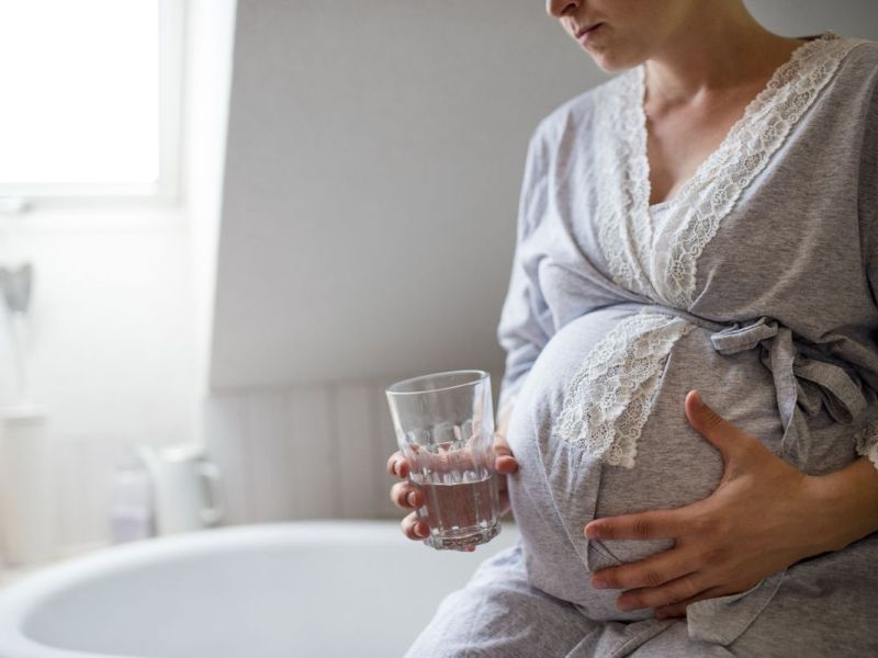Schwangere sitzt mit Wasserglas auf dem Badewannenrand und h&auml;lt ihren Bauch mit der anderen Hand.