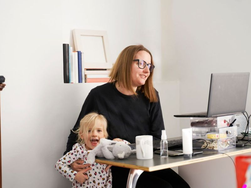 Berufst&auml;tuge Mutter sitzt vor dem Laptop, ihr Kleinkind neben sich.