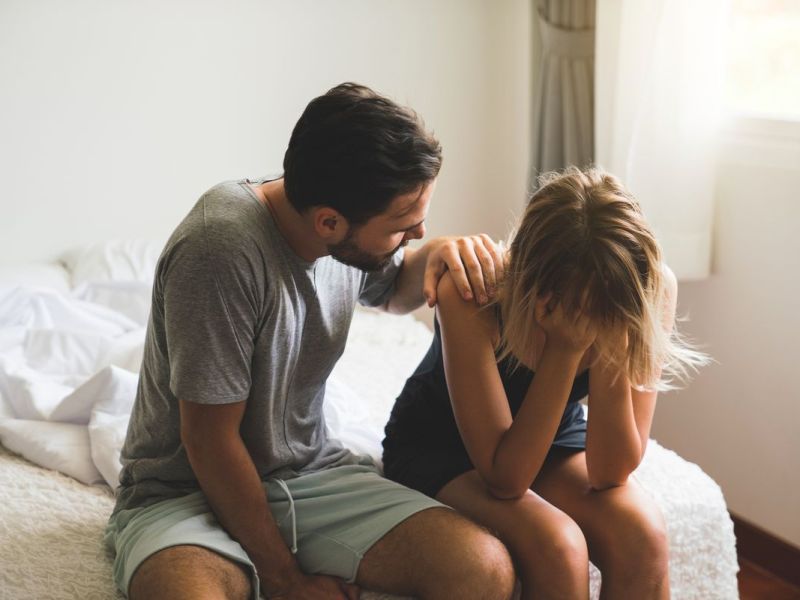 Frau sitzt auf dem Bett und hat die Hände unglücklich vors Gesicht geschlagen. Ein Mann beugt sich über sie und legt die Hand auf ihre Schulter. Das Paar wirkt unglücklich.