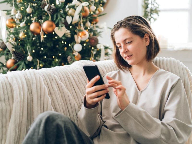 Frau sitzt mit einem Handy auf der Couch vor dem Weihnachtsbaum.