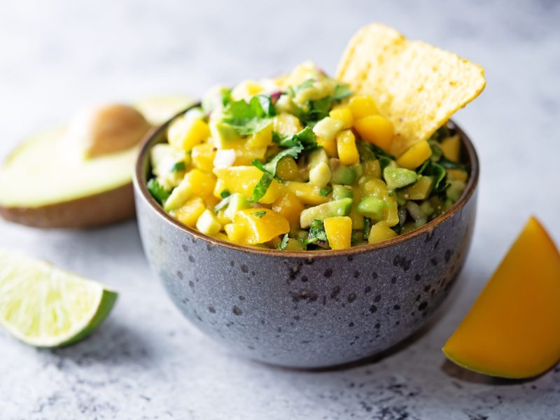 Eine Sch&uuml;ssel Mango-Avocado-Salsa mit eine Nacho-Chip, daneben je eine angeschnittene Limette, Avocado und Mango.