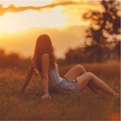 Frau im Sommer auf einem Feld, die sitzt und sich mit den H&auml;nden abst&uuml;tzt, w&auml;hrend sie den Sonnenuntergang anschaut