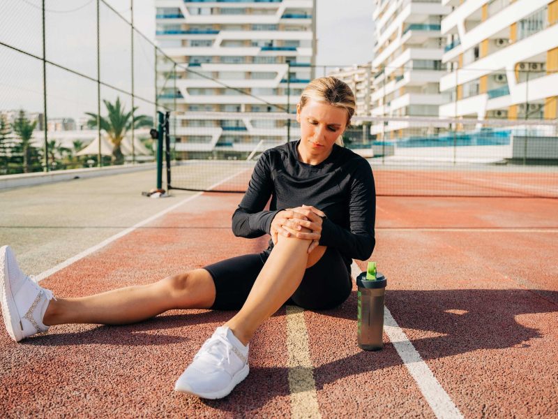 Athletische Frau h&auml;lt sich das schmerzende Knie auf einem Tennisplatz