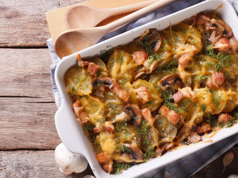 Eine wei&szlig;e Auflaufform auf einem hellen Holztisch mit dem Gratin und einem Bund Kr&auml;uter sowie Pilzen und Knoblauchknollen.
