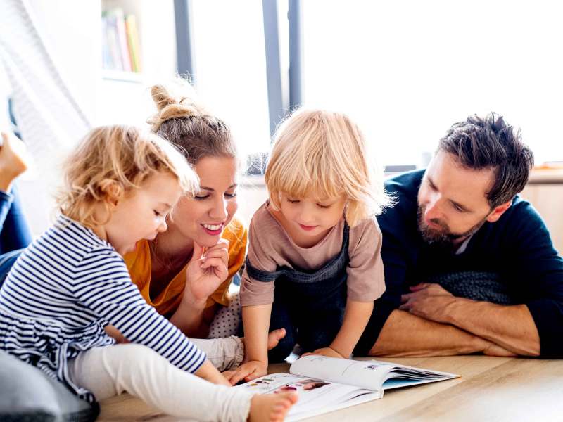 Mutter, Vater und zwei Kinder liegen auf dem Fu&szlig;boden und bl&auml;ttern gemeinsam in einem Buch.
