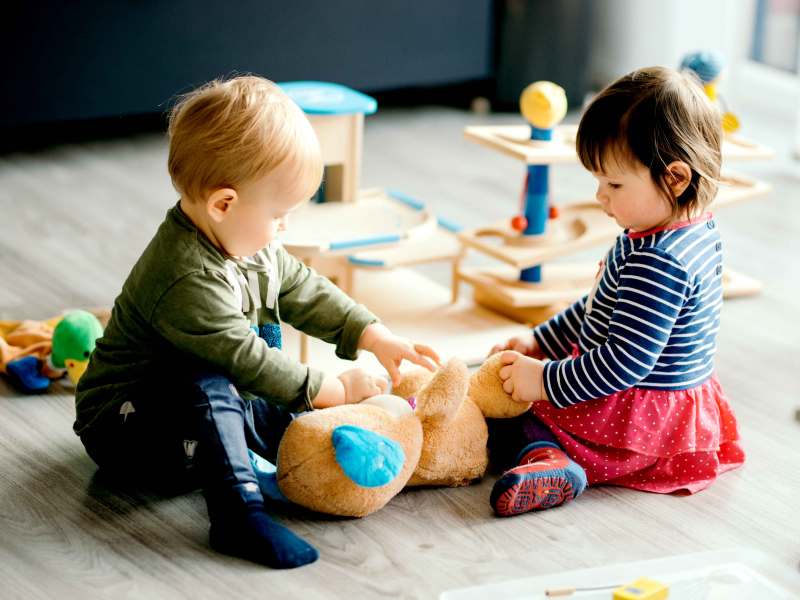 Zwei Kleinkinder sitzen auf dem Fu&szlig;boden und spielen mit einem gro&szlig;en Pl&uuml;schteddy.