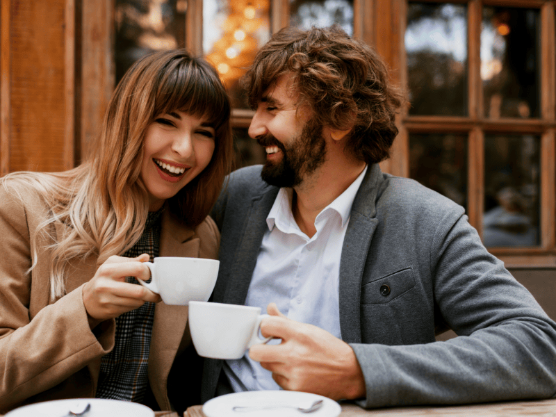 Eine junge Frau mit braunen Haaren und ein b&auml;rtiger Mann lachen sich an, w&auml;hrend sie drau&szlig;en vor einem Caf&eacute; Kaffee trinken.