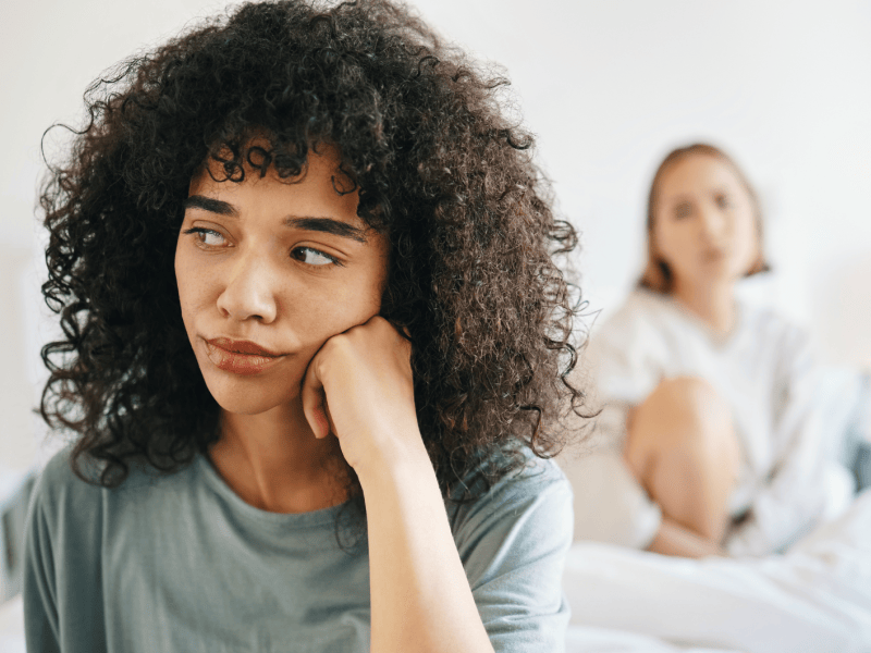 Frau, die nachdenklich zur Seite schaut, w&auml;hrend im Hintergrund eine andere Frau auf sie blickt und auf einem Bett sitzt.