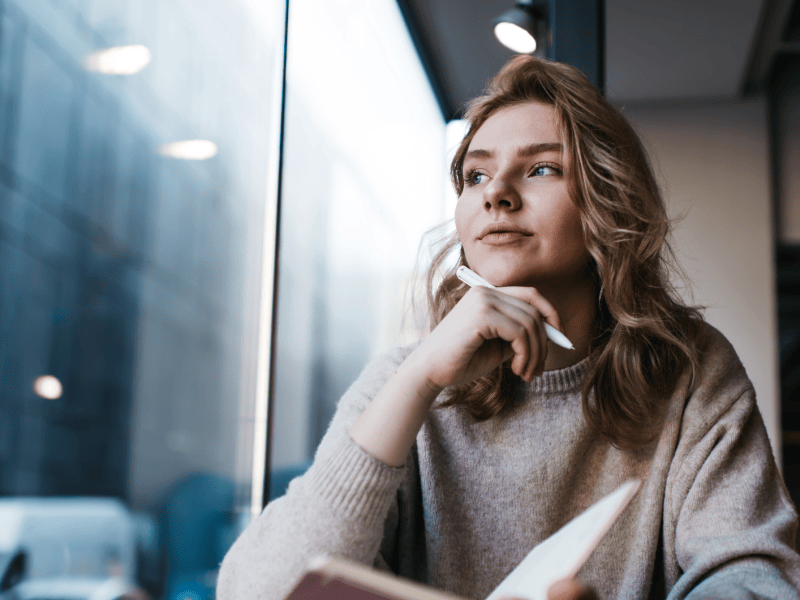 Frau vor einem Fenster in einem Caf&eacute;, die ein Buch in der Hand h&auml;lt und einen Stift an ihr Kinn h&auml;lt