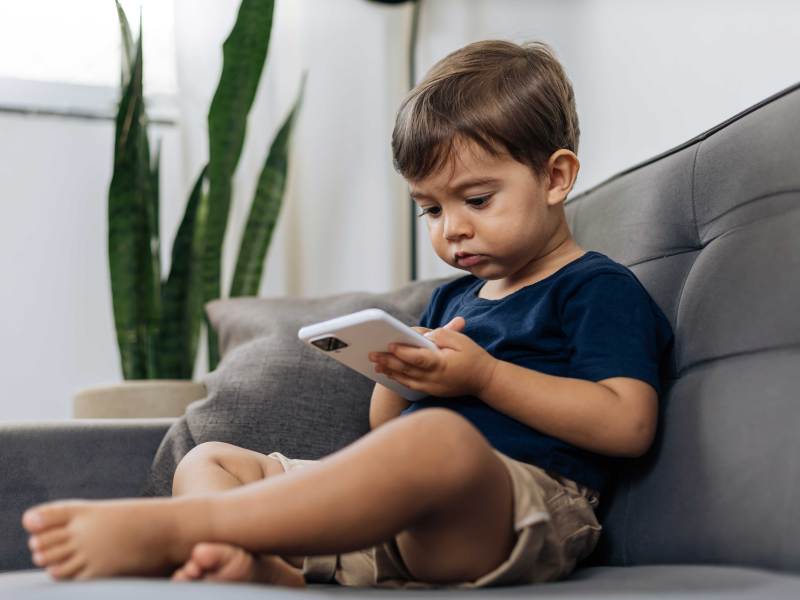 Kleiner, ca. 3-j&auml;hriger Junge sitzt mit einem Smartphone auf dem Sofa.