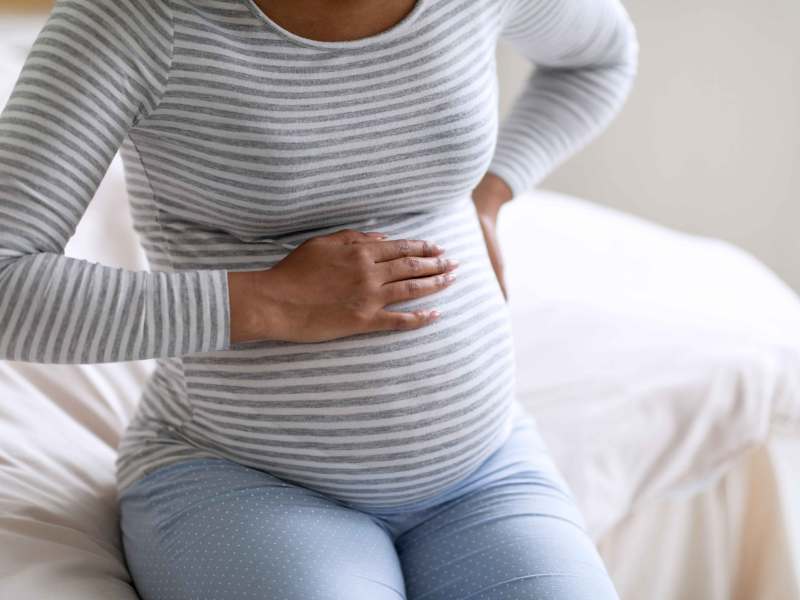 Schwangere Frau sitzt auf dem Bett und h&auml;lt sich sowohl den Bauch als auch den R&uuml;cken.