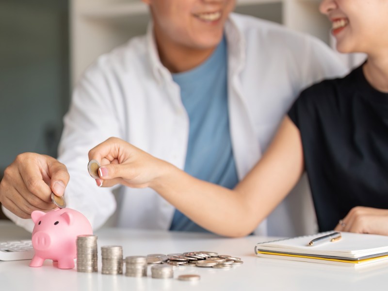 Paar wirft Geld in ein Sparschwein. Daneben sind M&uuml;nzen in kleinen T&uuml;rmen angeh&auml;uft.