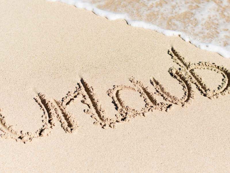 Das Wort &bdquo;Urlaub&ldquo; ist in den feinen Sand eines Strandes geschrieben, w&auml;hrend sanfte Wellen ans Ufer rollen.