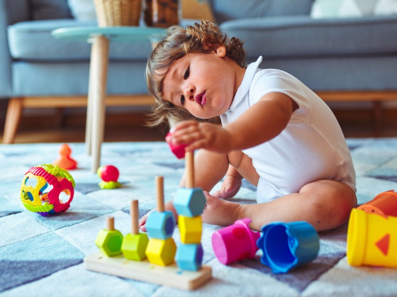 Kind, ca. zwei Jahre, sitzt auf dem Fu&szlig;boden des Wohnzimmers und spielt konzentriert mit Stapelspielzeug.