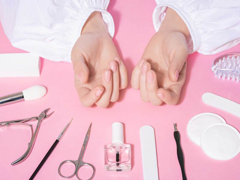 Nagelstudio Nahaufnahme von weiblichen gepflegten H&auml;nden und frisch manik&uuml;rten Fingern&auml;geln mit rosa Hintergrund. Sch&ouml;nheit und Pflege-Industrie-Konzept mit Manik&uuml;re-Tools, wie Schere, Zange und Pinzette.