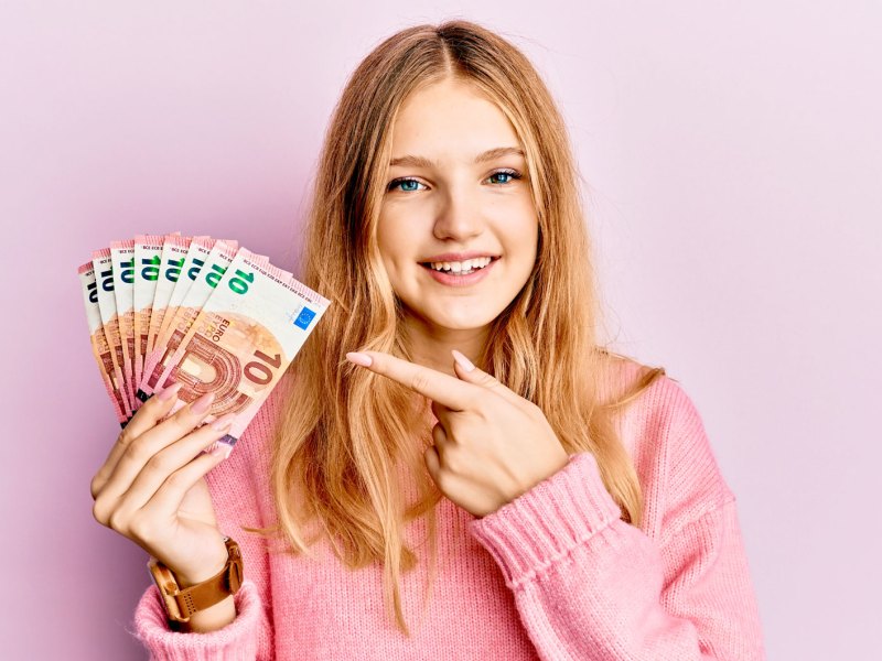 M&auml;dchen vor rosa Hintergrund h&auml;lt ein paar Zehn Euro Scheine in die Kamera und zeigt darauf.