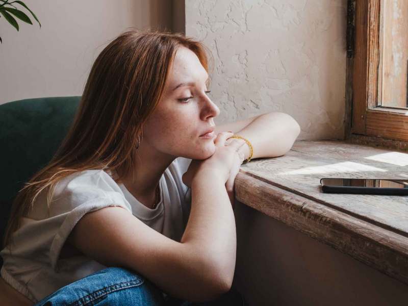 Frau sitzt am Fenster und guckt ungl&uuml;cklich auf ihr Handy, das auf der Fensterbank vor ihr liegt.