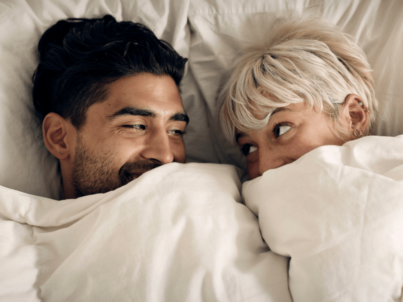 Ein dunkelhaariger Mann und eine blondhaarige Frau schauen sich l&auml;chelnd unter einer wei&szlig;en Bettdecke verliebt in die Augen und lachen.