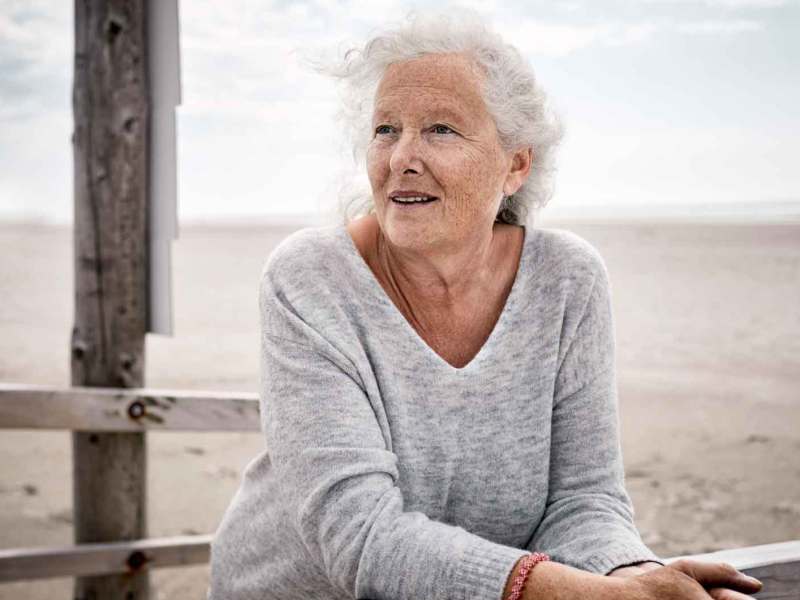 &Auml;ltere Frau steht am Strand an einen Zaun angelehnt.