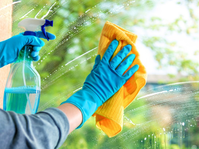 Person putzt Fenster, h&auml;lt gelbes Tuch in einer Hand und Spr&uuml;hflasche mit Reiniger in anderer.