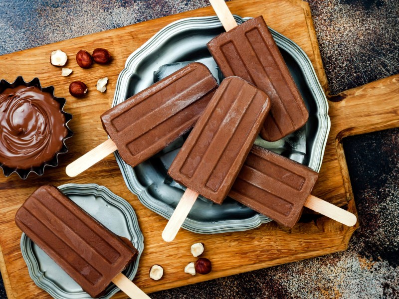 Schokoladen-Stieleis auf einem Holzbrett, daneben eine kleine Sch&uuml;ssel mit Schokocreme.