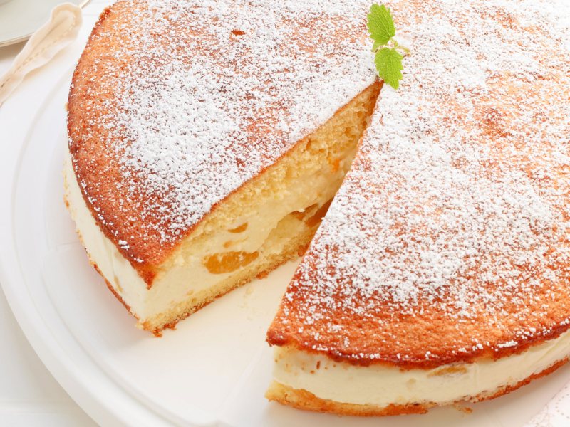 Luftige Schmandtorte mit Pfirsich-St&uuml;ckchen in der Creme, angeschnitten auf einem wei&szlig;en Teller.