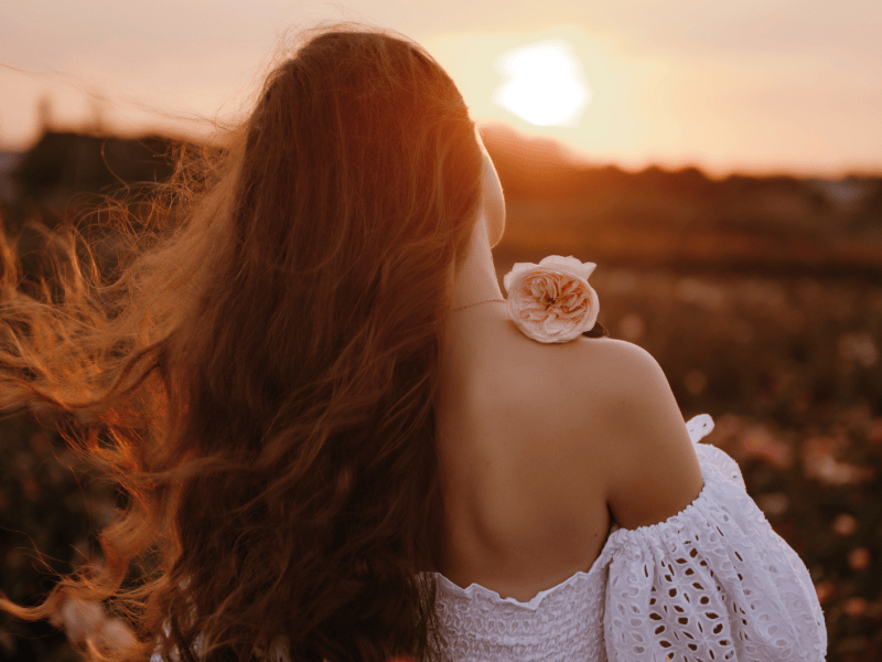 Frau auf einem Feld in einem wei&szlig;en Kleid mit Blume in der Hand, die sich mit dem R&uuml;cken zur Kamera dreht.