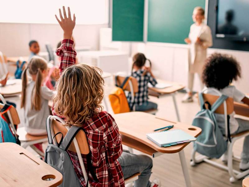 Blick in ein Klassenzimmer. Die Kinder heben zum Teil die H&auml;nde, die Lehrerin steht vorn vor der Tafel, mit dem Blick ins Klassenzimmer.