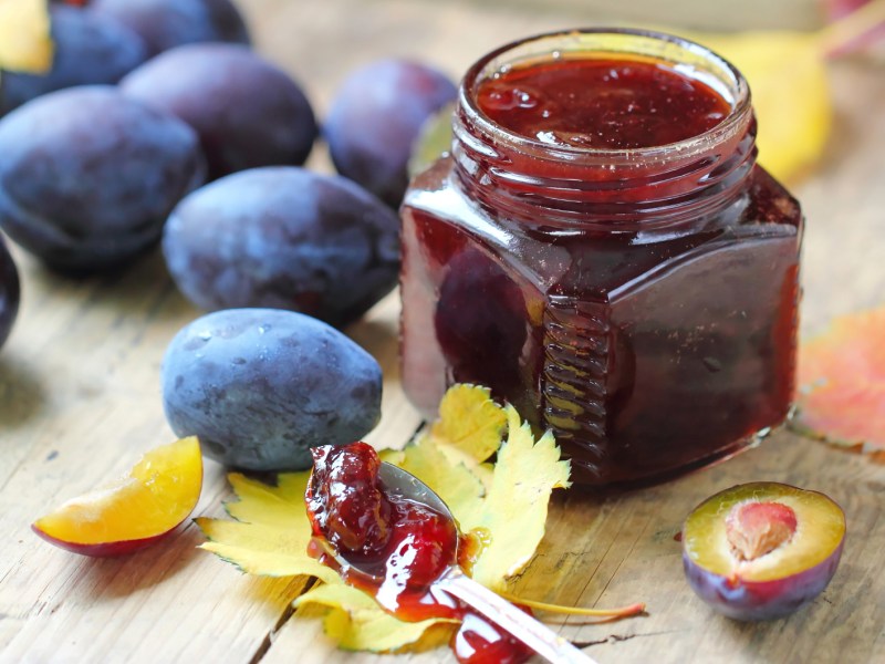 Pflaumenmarmelade in einem Weckglas, daneben liegen frische Pflaumen und Herbstbl&auml;tter.