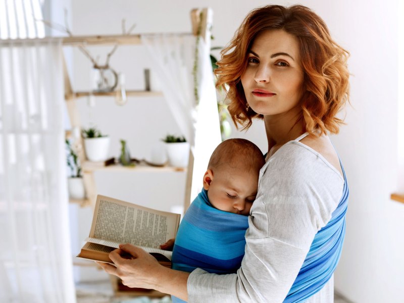 Portr&auml;t einer zufriedenen Mutter, die ihr Kind in einem Tragetuch vor der Brust tr&auml;gt und ein Buch in den H&auml;nden h&auml;lt.