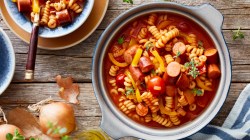 Wurstgulasch mit Nudeln und Paprika in großem Topf auf Holztisch.
