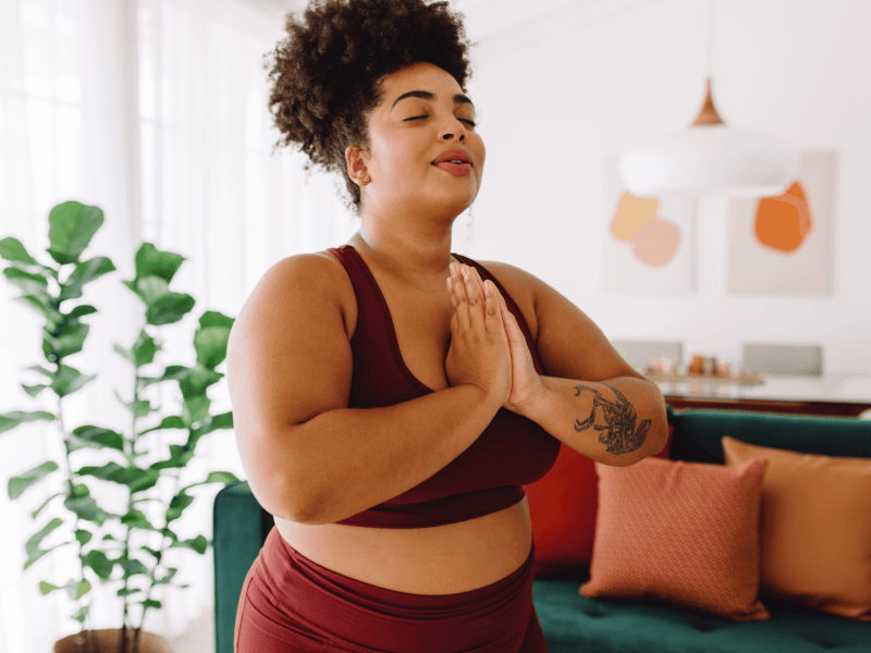 Frau vor Couch, die Yoga macht und die Hände vor der Brust zusammenlegt