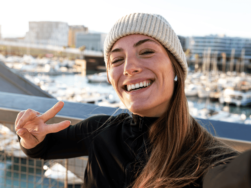 Fr&ouml;hliche Frau in schwarzer Jacke und beige Strickm&uuml;tze zeigt ein Peace-Zeichen, w&auml;hrend sie ein Selfie vor einem Yachthafen aufnimmt.