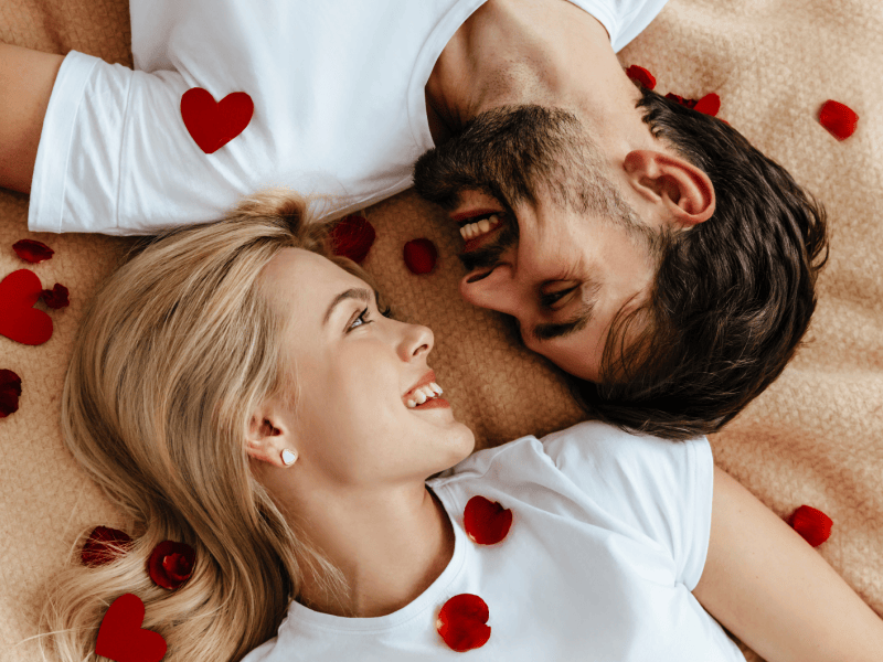 Ein Mann und eine Frau, die auf einer Decke gemeinsam liegen und sich zwischen Rosenbl&auml;ttern verliebt anschauen.