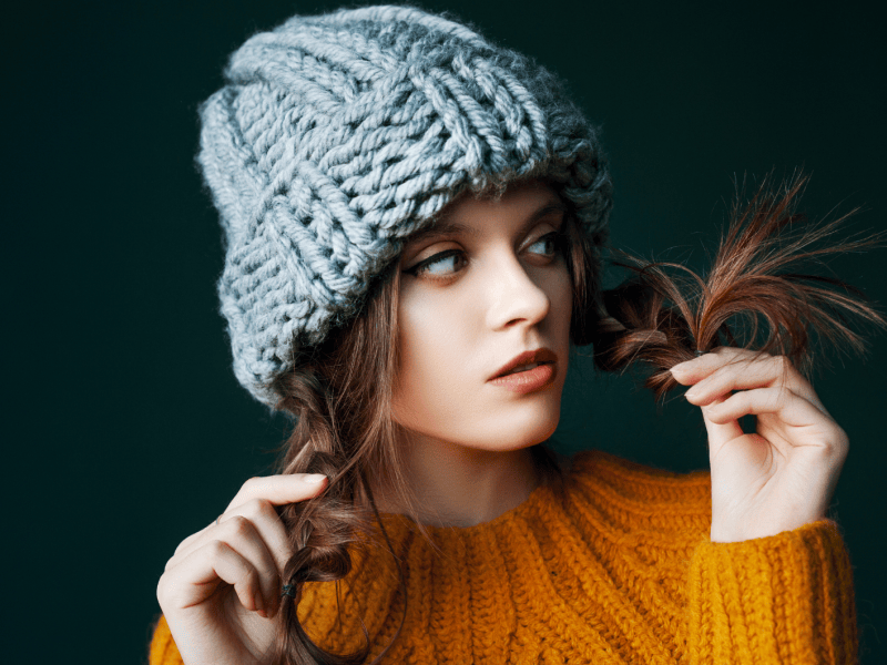 Eine junge Frau in einem gelben Strickpullover und einer grob gestrickten grauen M&uuml;tze. Sie h&auml;lt spielerisch ihre braunen Haarspitzen mit den H&auml;nden, w&auml;hrend sie seitlich schaut. Der Hintergrund ist dunkelgr&uuml;n.