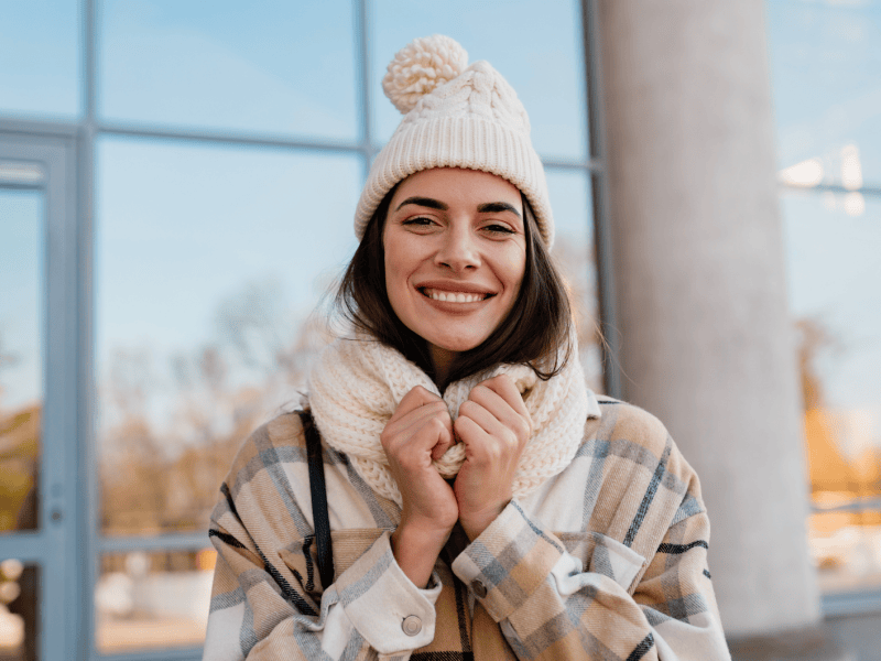 Fr&ouml;hliche Frau mit beigem Winteroutfit h&auml;lt Schal fest, sonniger Tag, reflektierende Fenster im Hintergrund.