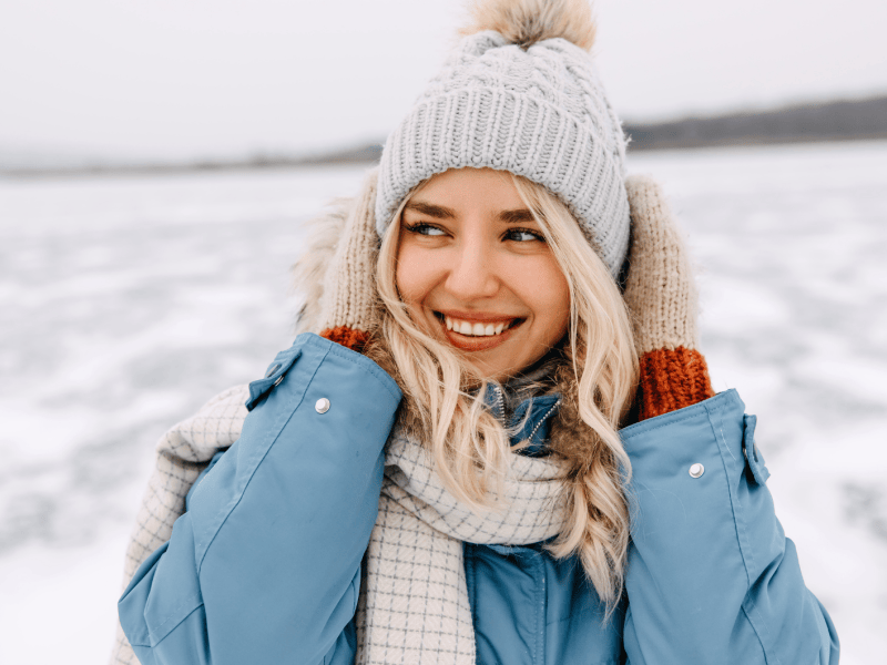 Frau im Schnee, mit Winterjacke, M&uuml;tze und Schal, die sich an die Ohren packt und zur Seite l&auml;chelt.