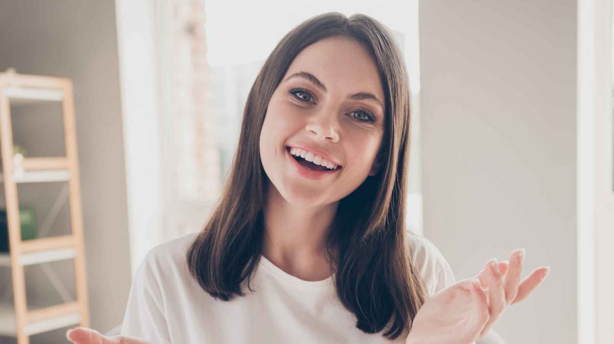 Eine junge Frau mit schulterlangem braunem Haar und weißem T-Shirt lächelt freundlich in die Kamera, sitzend in einem hellen Raum.