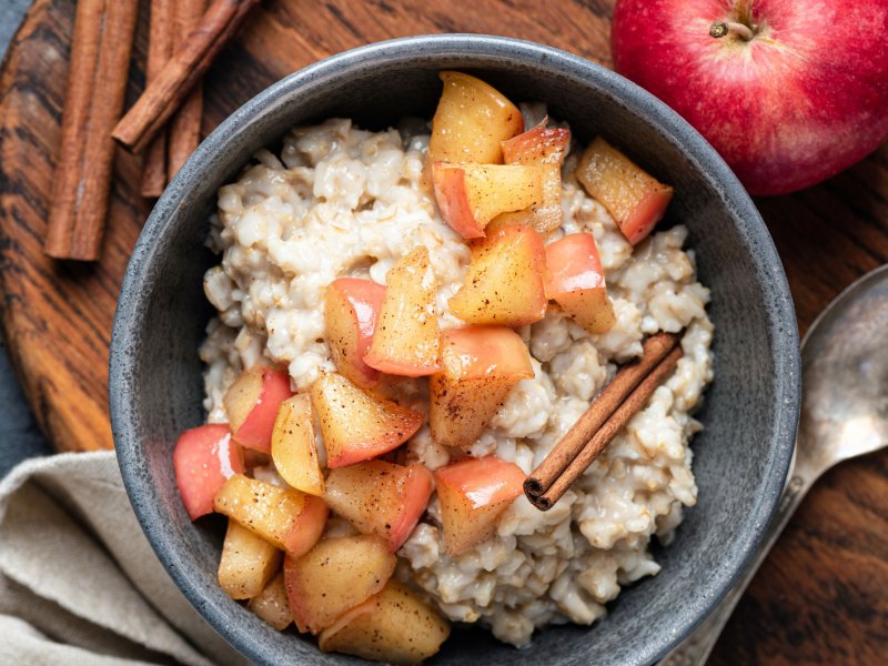 Apfel-Zimt-Porridge in einer schwarzen Sch&uuml;ssel auf einem Holzbrett. Ein Apfel, ein L&ouml;ffel & Zimtstangen liegen daneben.