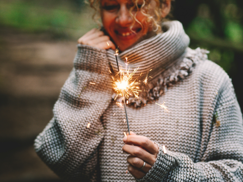 Eine lachende junge Frau mit lockigem blondem Haar h&auml;lt eine brennende Wunderkerze, tr&auml;gt einen groben Wollpullover im Wald.