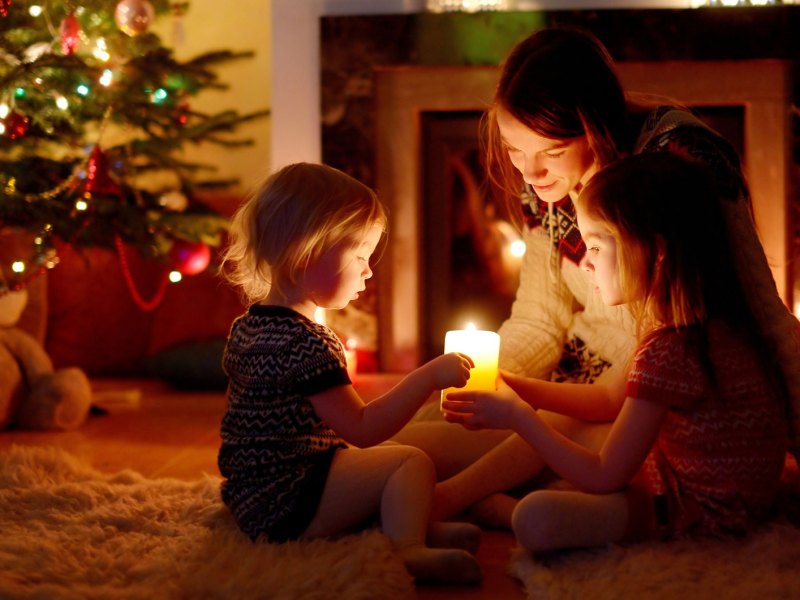 Mutter und T&ouml;chter sitzen auf dem Fu&szlig;boden neben ihrem Weihnachtsbaum und halten gemeinsam eine einzige Kerze.