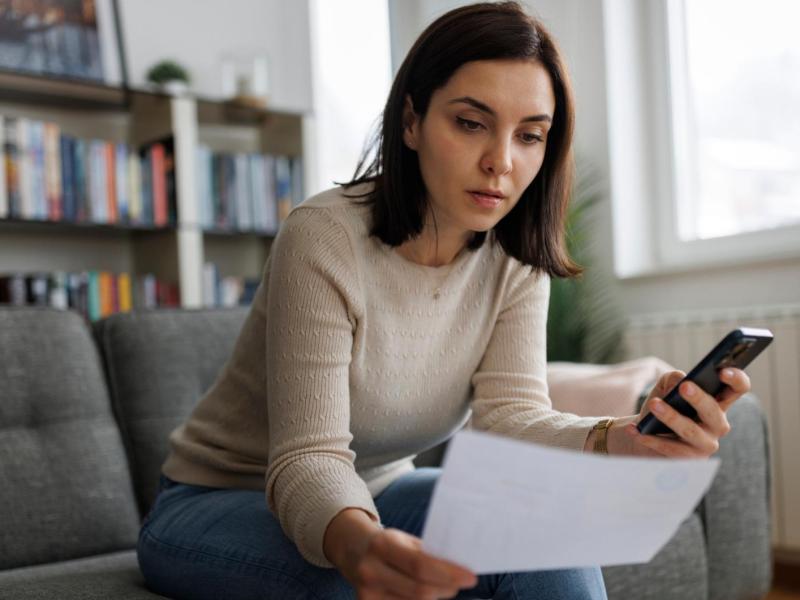 Eine junge Frau mit schulterlangem braunem Haar sitzt nachdenklich auf einem grauen Sofa, h&auml;lt ein Dokument und schaut auf ihr Smartphone.
