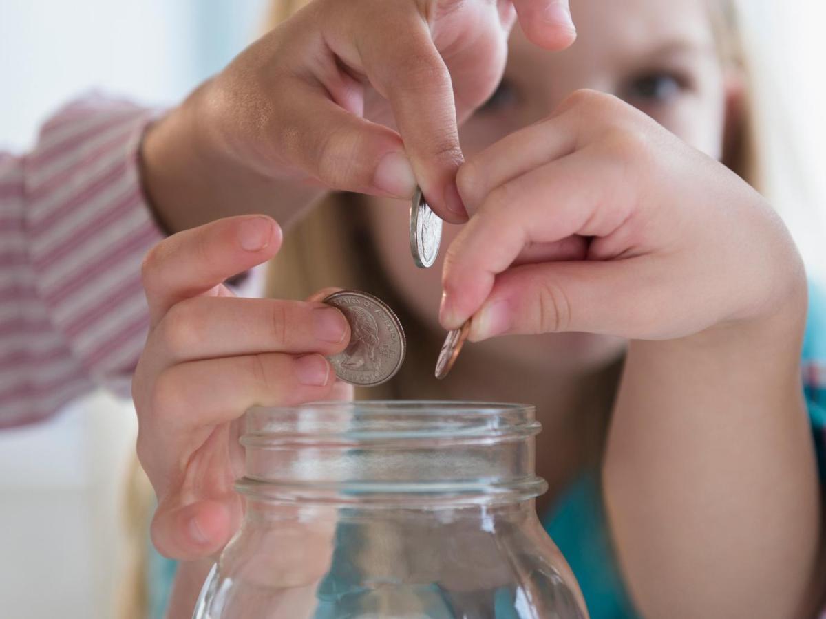Drei Kinderh&auml;nde werfen konzentriert M&uuml;nzen in ein gro&szlig;es, leeres Einmachglas vor unscharfem, hellem Hintergrund.